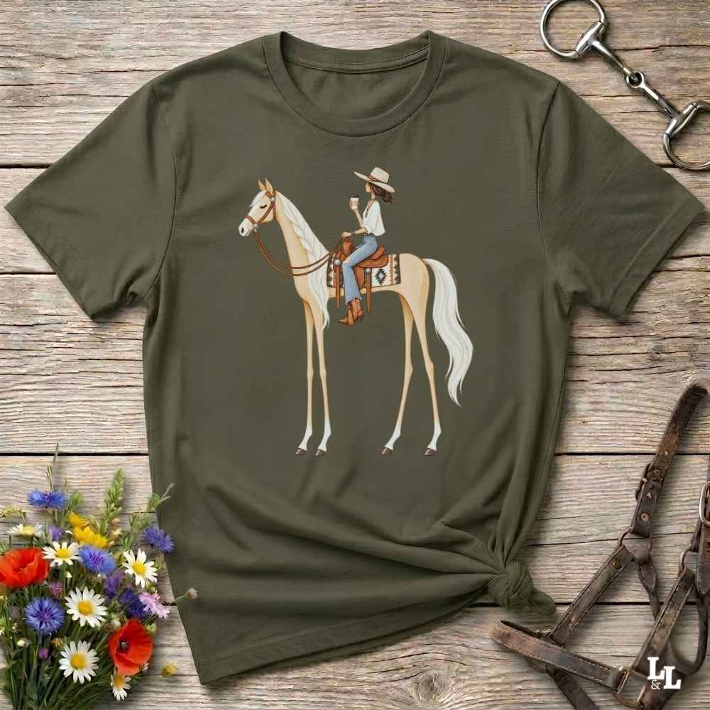 An olive green short-sleeve t-shirt with a graphic of a woman riding a light-colored horse, styled in a flat-lay on a rustic wooden background with flowers and horse tack.