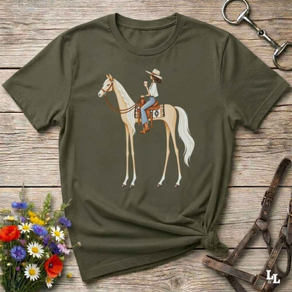 An olive green short-sleeve t-shirt with a graphic of a woman riding a light-colored horse, styled in a flat-lay on a rustic wooden background with flowers and horse tack.