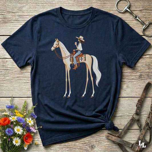 A navy blue short-sleeve t-shirt with a graphic of a woman riding a light-colored horse, styled in a flat-lay on a rustic wooden background with equestrian accessories and flowers.