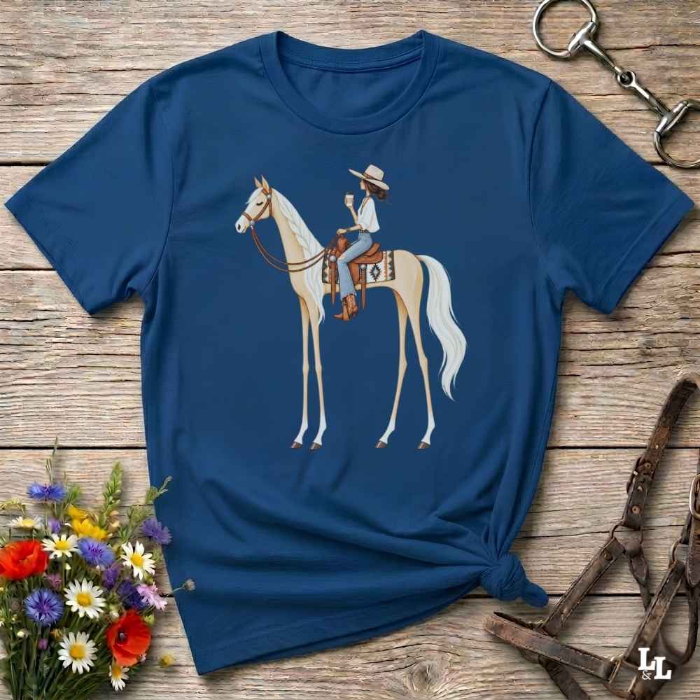 A navy blue short-sleeve t-shirt with a graphic of a cowboy riding a light-colored horse, styled in a flat-lay on a rustic wooden surface with equestrian accessories and flowers.