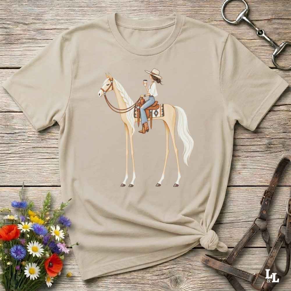 A beige graphic t-shirt featuring an illustration of a woman riding a light-colored horse, styled in a flat-lay on a rustic wooden background with equestrian accessories and flowers.