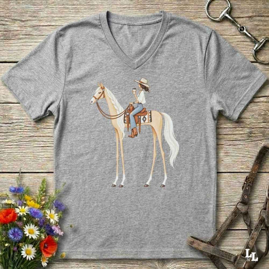 A heather gray V-neck t-shirt featuring a graphic of a woman riding a light-colored horse, styled in a flat-lay on a rustic wooden background with equestrian accessories and flowers.