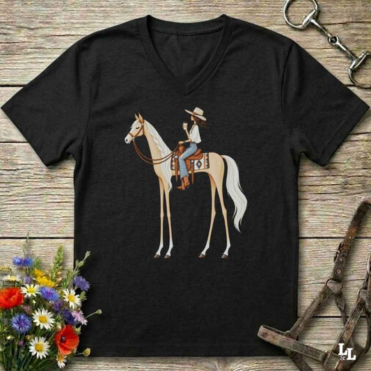 A black V-neck t-shirt featuring a graphic illustration of a woman riding a light-colored horse, displayed on a rustic wooden background with equestrian accessories and flowers.
