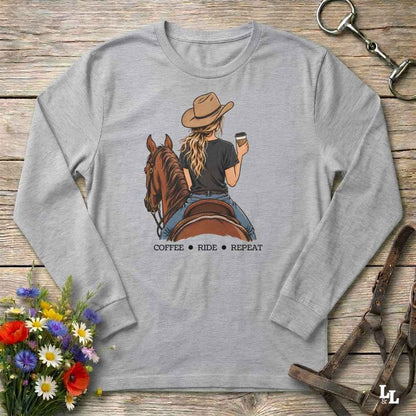 A heather gray long-sleeve t-shirt with a graphic of a woman riding a brown horse while holding a coffee cup, with the text 'COFFEE RIDE REPEAT' underneath. The shirt is displayed flat on a rustic wooden background with decorative flowers and leather tack.
