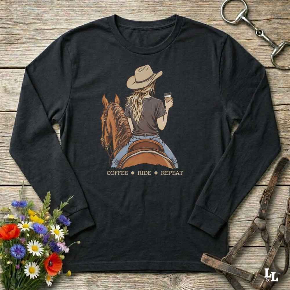A black long-sleeve t-shirt with a graphic of a woman riding a brown horse while holding a cup of coffee, with the text 'COFFEE RIDE REPEAT' underneath. The shirt is laid flat on a rustic wooden background with decorative props including a bouquet of flowers, a horse bit, and a leather bridle.