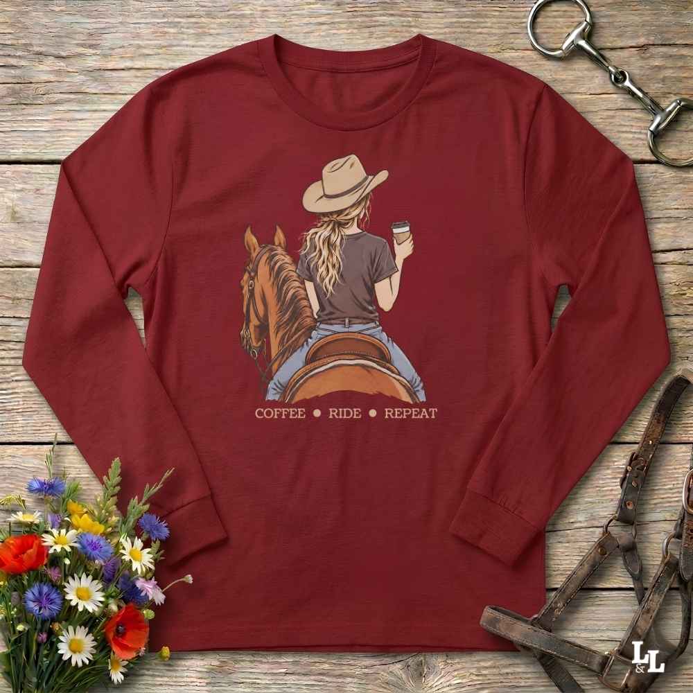 A long-sleeve crewneck t-shirt in maroon red, laid flat on a rustic wooden surface. The shirt features a graphic illustration of a woman in a cowboy hat riding a horse while holding a cup of coffee, with the text 'COFFEE RIDE REPEAT' printed below.