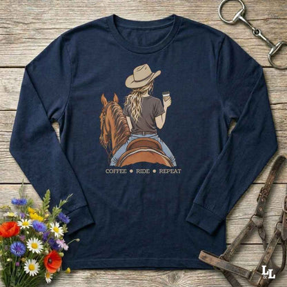 A navy blue long-sleeve t-shirt with a graphic of a woman riding a brown horse while holding a cup of coffee, with the text 'COFFEE RIDE REPEAT' underneath. The shirt is displayed in a flat-lay arrangement on a rustic wooden background, surrounded by decorative elements including a bouquet of flowers, a horse bit, and a leather bridle.