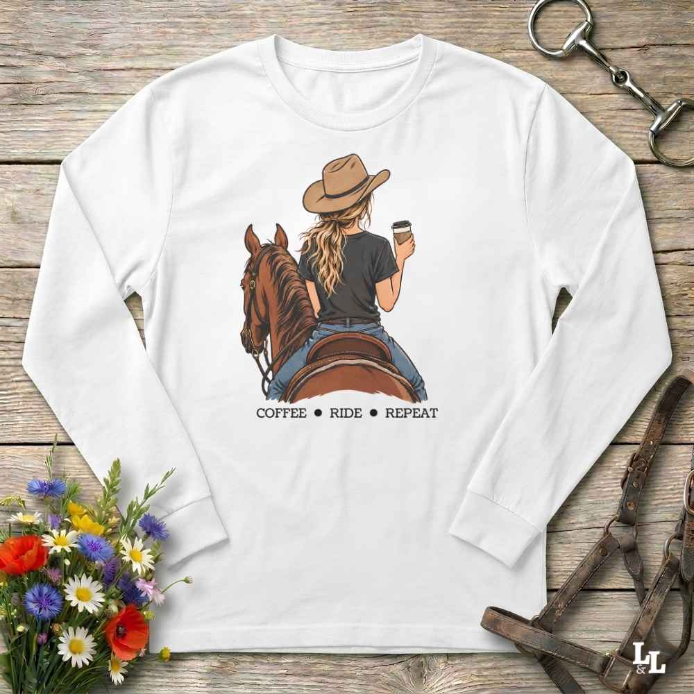 A white long-sleeve t-shirt with a graphic of a woman on a brown horse holding a coffee cup, with the text 'COFFEE RIDE REPEAT' underneath. The shirt is laid flat on a rustic wooden background with equestrian accessories and flowers.