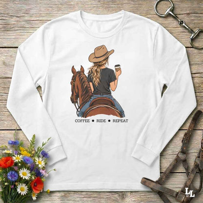 A white long-sleeve t-shirt with a graphic of a woman on a brown horse holding a coffee cup, with the text 'COFFEE RIDE REPEAT' underneath. The shirt is laid flat on a rustic wooden background with equestrian accessories and flowers.