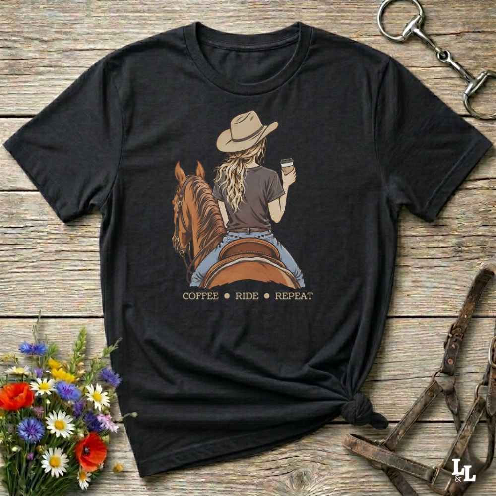 A black short-sleeve graphic T-shirt laid flat on a rustic wooden surface, styled with flowers, a horse bit, and leather tack. The shirt features a printed illustration of a woman in a cowboy hat riding a brown horse while holding a coffee cup, with the words 'COFFEE RIDE REPEAT' below.