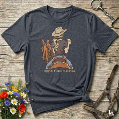 A dark heather gray graphic t-shirt laid flat on a rustic wooden surface, styled with equestrian and floral props. The graphic shows a woman in a cowboy hat riding a brown horse and holding a cup of coffee, with the text 'COFFEE RIDE REPEAT' underneath.