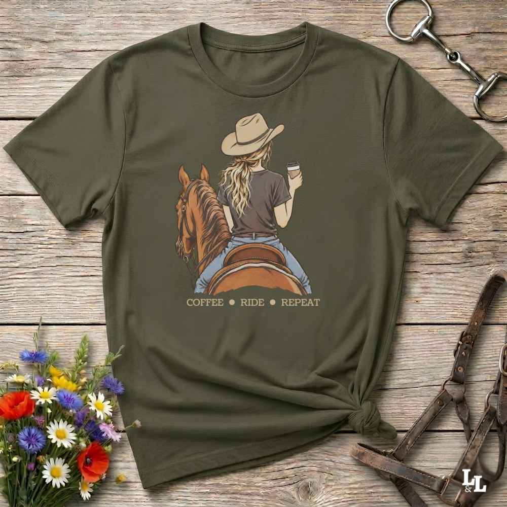 An olive green graphic t-shirt laid flat on a rustic wooden surface, styled with a horse and bit in the top right and a small bouquet of flowers in the bottom left. The t-shirt features a graphic of a woman in a cowboy hat riding a brown horse while holding a cup of coffee, with the text 'COFFEE RIDE REPEAT' printed below.