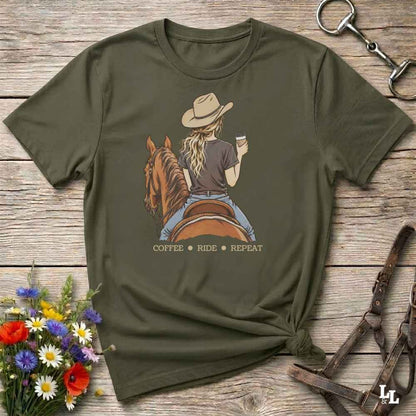 An olive green graphic t-shirt laid flat on a rustic wooden surface, styled with a horse and bit in the top right and a small bouquet of flowers in the bottom left. The t-shirt features a graphic of a woman in a cowboy hat riding a brown horse while holding a cup of coffee, with the text 'COFFEE RIDE REPEAT' printed below.