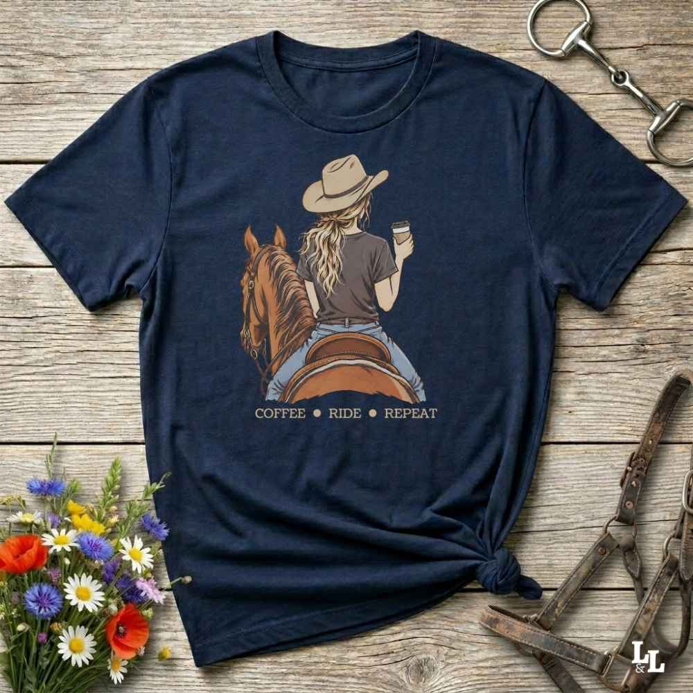 A navy blue graphic t-shirt laid flat on a rustic wooden surface, styled with equestrian and floral props. The shirt features a graphic of a woman in a cowboy hat riding a brown horse while holding a cup of coffee, with the text 'COFFEE RIDE REPEAT' printed below.