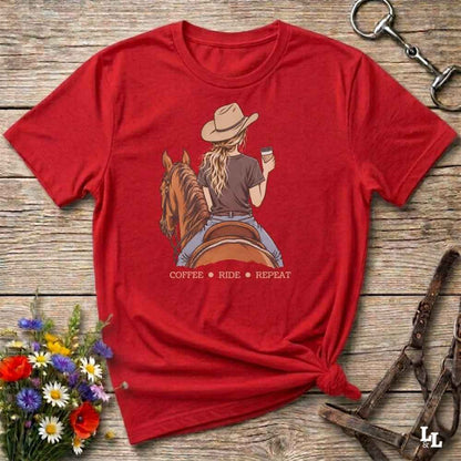 A red short-sleeve graphic T-shirt laid flat on a rustic wooden surface, styled with flowers, a horse bit, and a leather bridle. The shirt features a printed illustration of a woman in a cowboy hat riding a horse while holding a coffee cup, with the text 'COFFEE RIDE REPEAT' below.