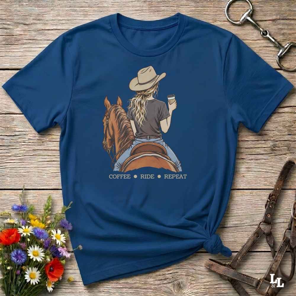 A navy blue graphic t-shirt laid flat on a rustic wooden surface, styled with a cowboy hat, horse, and floral props. The graphic shows a woman riding a brown horse while holding a coffee cup, with the text 'COFFEE RIDE REPEAT' underneath.