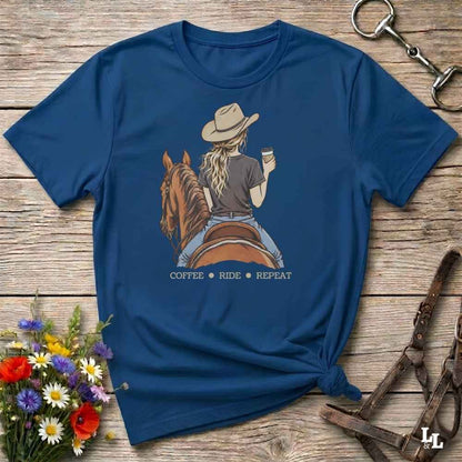 A navy blue graphic t-shirt laid flat on a rustic wooden surface, styled with a cowboy hat, horse, and floral props. The graphic shows a woman riding a brown horse while holding a coffee cup, with the text 'COFFEE RIDE REPEAT' underneath.