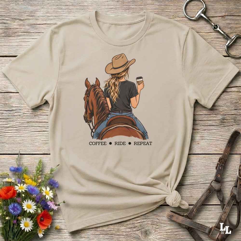 A beige graphic t-shirt featuring an illustration of a woman on a horse riding while holding a coffee cup, with the text 'COFFEE RIDE REPEAT' underneath. The t-shirt is styled in a flat-lay on a rustic wooden background with equestrian accessories and a small bouquet of wildflowers.