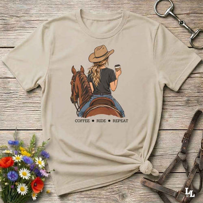 A beige graphic t-shirt featuring an illustration of a woman on a horse riding while holding a coffee cup, with the text 'COFFEE RIDE REPEAT' underneath. The t-shirt is styled in a flat-lay on a rustic wooden background with equestrian accessories and a small bouquet of wildflowers.