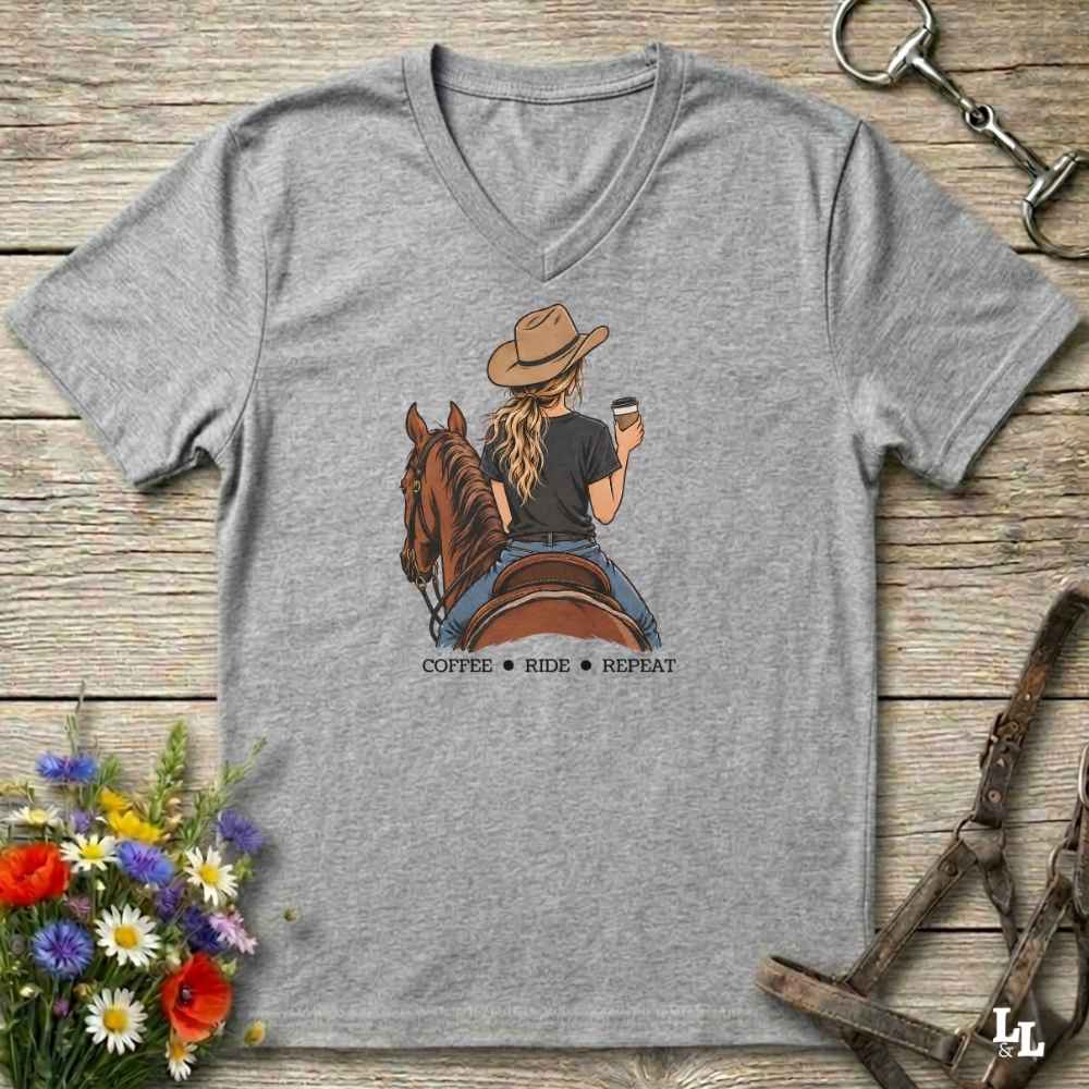 A heather gray V-neck t-shirt laid flat on a rustic wooden surface, featuring a graphic of a woman in a cowboy hat riding a brown horse while holding a coffee cup, with the text 'COFFEE RIDE REPEAT' underneath. The shirt is styled with equestrian and floral props.