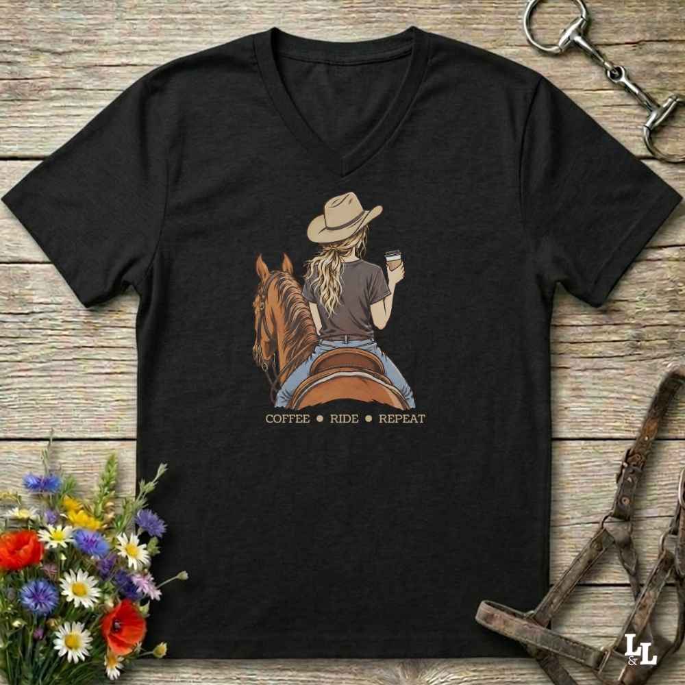 A black V-neck t-shirt laid flat on a rustic wooden surface, featuring a graphic of a woman in a cowboy hat riding a brown horse while holding a coffee cup, with the text 'COFFEE RIDE REPEAT' below. The shirt is styled with flowers, a horse bit, and a halter.