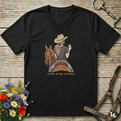 A black V-neck t-shirt laid flat on a rustic wooden surface, featuring a graphic of a woman in a cowboy hat riding a brown horse while holding a coffee cup, with the text 'COFFEE RIDE REPEAT' below. The shirt is styled with flowers, a horse bit, and a halter.