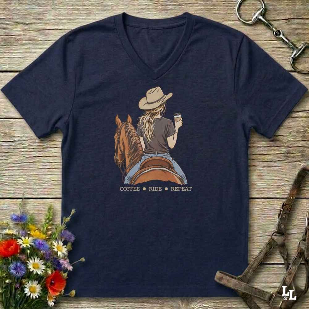 A navy blue V-neck t-shirt laid flat on a rustic wooden surface, featuring a graphic of a woman on a horse with the text 'COFFEE RIDE REPEAT'. The shirt is styled with equestrian props including a halter, tack, and a small bouquet of wildflowers.