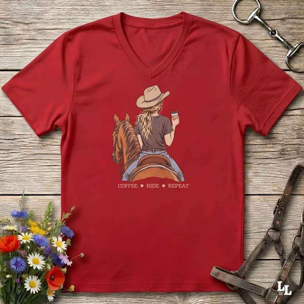 A red V-neck t-shirt laid flat on a rustic wooden surface, featuring a graphic of a woman in a cowboy hat riding a horse while holding a cup, with the text 'COFFEE RIDE REPEAT' underneath. The shirt is styled with equestrian accessories and a small bouquet of wildflowers.