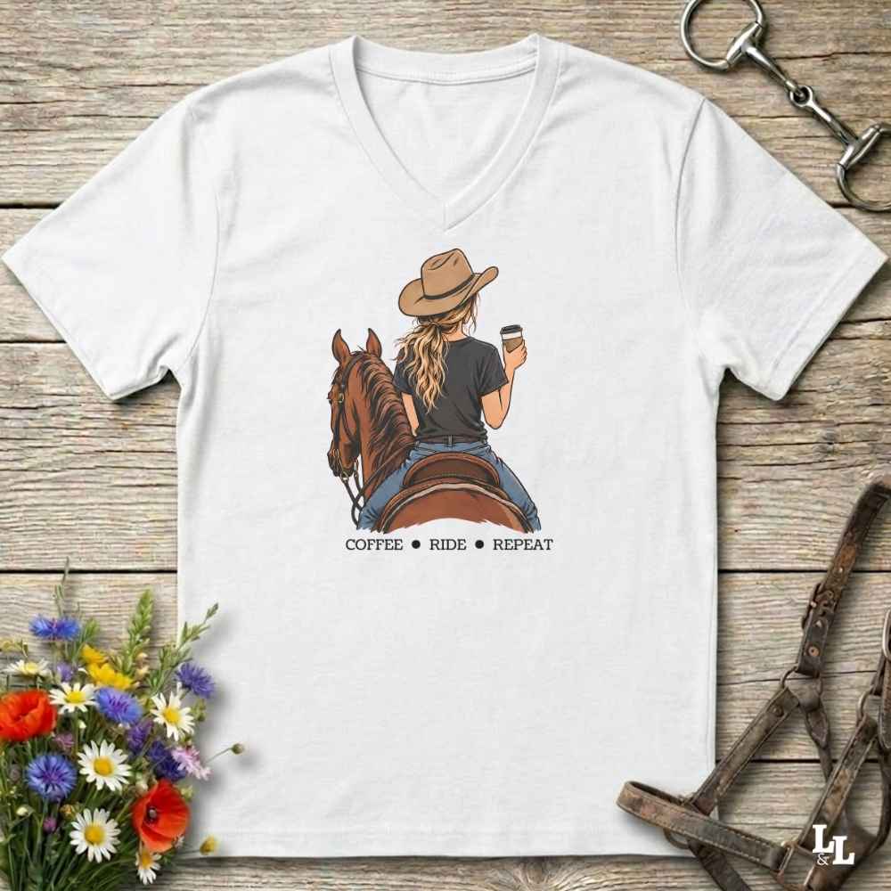 A white V-neck t-shirt laid flat on a rustic wooden surface, featuring a graphic of a woman in a cowboy hat riding a brown horse and holding a coffee cup, with the text 'COFFEE RIDE REPEAT' underneath. The shirt is styled with equestrian accessories (reins, bridle, and a horse bit) and colorful wildflowers.