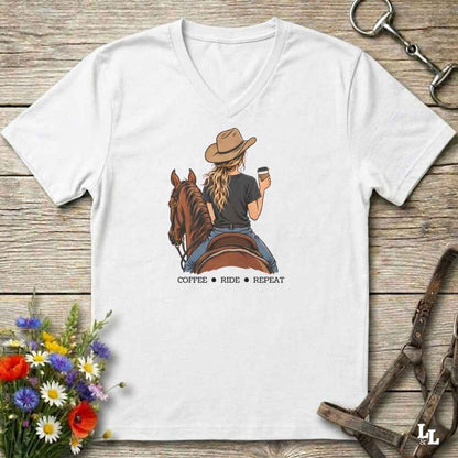 A white V-neck t-shirt laid flat on a rustic wooden surface, featuring a graphic of a woman in a cowboy hat riding a brown horse and holding a coffee cup, with the text 'COFFEE RIDE REPEAT' underneath. The shirt is styled with equestrian accessories (reins, bridle, and a horse bit) and colorful wildflowers.
