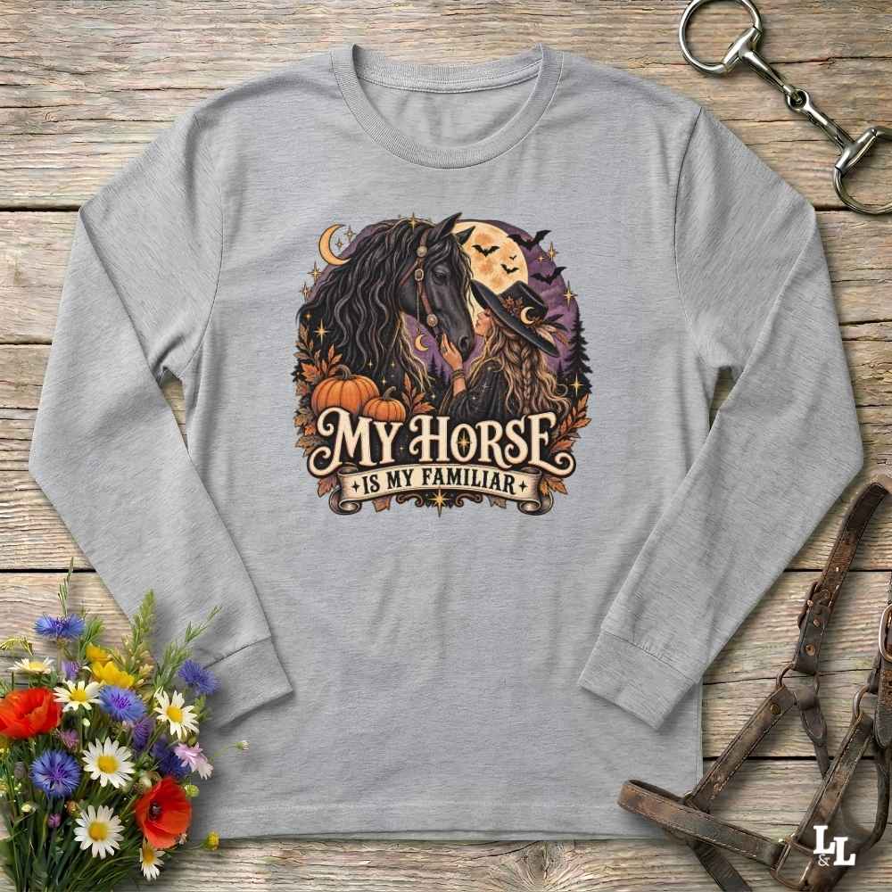 A light gray long-sleeve t-shirt with a graphic print of a witch horse and the text 'MY HORSE IS MY FAMILIAR'. The shirt is displayed in a flat-lay arrangement on a rustic wooden background, styled with a bouquet of flowers, a horse bit, and a leather bridle.