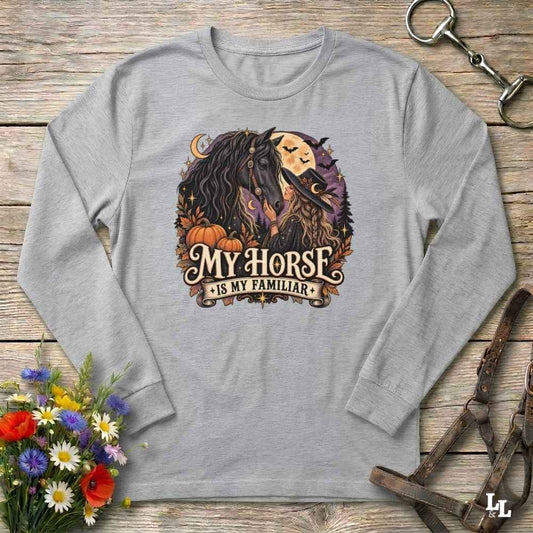 A light gray long-sleeve t-shirt with a graphic print of a witch horse and the text 'MY HORSE IS MY FAMILIAR'. The shirt is displayed in a flat-lay arrangement on a rustic wooden background, styled with a bouquet of flowers, a horse bit, and a leather bridle.
