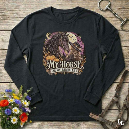 A black long-sleeve t-shirt with a graphic print of a horse and a skull under a full moon, surrounded by pumpkins and the text 'MY HORSE IS MY FAMILIAR'. The shirt is displayed flat on a rustic wooden background with decorative props including a bouquet of flowers, a horse bit, and a leather bridle.