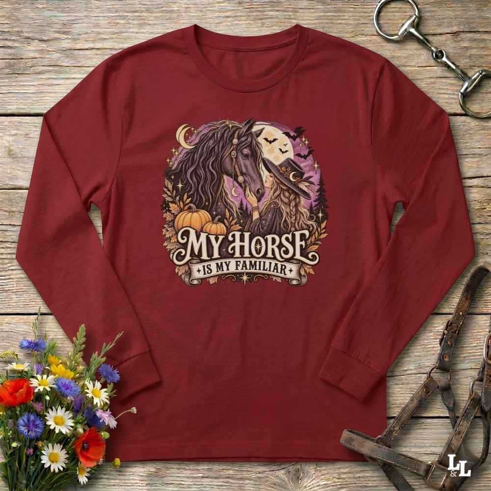 A long-sleeve crewneck t-shirt in maroon red, laid flat on a rustic wooden background. The shirt features a graphic print of a horse and a spooky cat under a full moon with pumpkins, and the text 'MY HORSE IS MY FAMILIAR'.