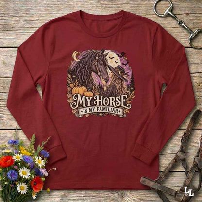 A long-sleeve crewneck t-shirt in maroon red, laid flat on a rustic wooden background. The shirt features a graphic print of a horse and a spooky cat under a full moon with pumpkins, and the text 'MY HORSE IS MY FAMILIAR'.