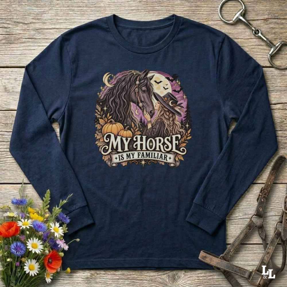 A navy blue long-sleeve t-shirt with a graphic of a horse and a skull under a full moon, surrounded by pumpkins and the text 'MY HORSE IS MY FAMILIAR'. The shirt is displayed flat on a rustic wooden background with decorative flowers and horse tack.