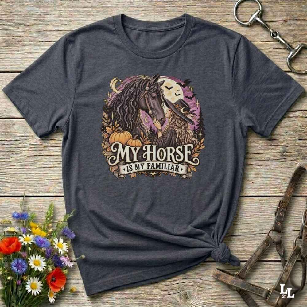 A dark charcoal gray graphic t-shirt laid flat on a rustic wooden surface, styled with a horse halter, a bouquet of wildflowers, and a small horse bit. The t-shirt features a graphic of a horse and moon with the text 'MY HORSE IS MY FAMILIAR'.