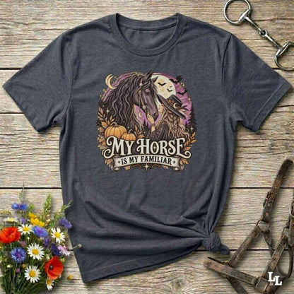 A dark charcoal gray graphic t-shirt laid flat on a rustic wooden surface, styled with a horse halter, a bouquet of wildflowers, and a small horse bit. The t-shirt features a graphic of a horse and moon with the text 'MY HORSE IS MY FAMILIAR'.