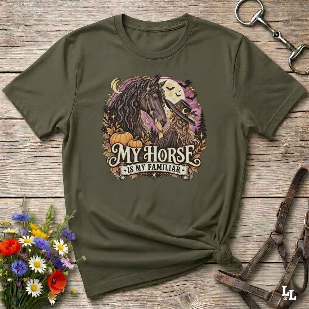 An olive green graphic t-shirt laid flat on a rustic wooden surface, styled with a horse halter, a small bouquet of wildflowers, and a horse bridle. The t-shirt features a circular graphic of a horse and a skull under a full moon with the text 'MY HORSE IS MY FAMILIAR'.