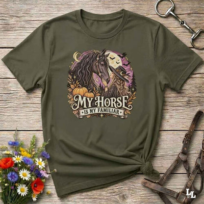 An olive green graphic t-shirt laid flat on a rustic wooden surface, styled with a horse halter, a small bouquet of wildflowers, and a horse bridle. The t-shirt features a circular graphic of a horse and a skull under a full moon with the text 'MY HORSE IS MY FAMILIAR'.