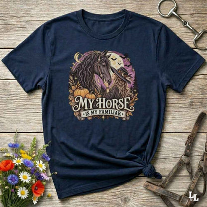 A navy blue graphic t-shirt laid flat on a rustic wooden surface, styled with a small bunch of wildflowers on the bottom left and equestrian tack props on the right. The t-shirt features a circular graphic of a dark-haired horse and a white moon with the text 'MY HORSE IS MY FAMILIAR' in a vintage font.