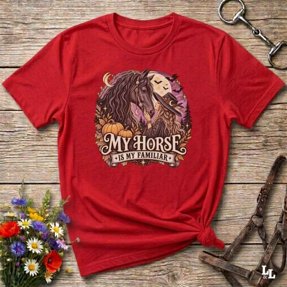 A red short-sleeve graphic T-shirt laid flat on a rustic wooden surface, styled with flowers, a horse bit, and a leather bridle. The shirt features a large front print of a horse and rider with pumpkins and the text 'MY HORSE IS MY FAMILIAR'.