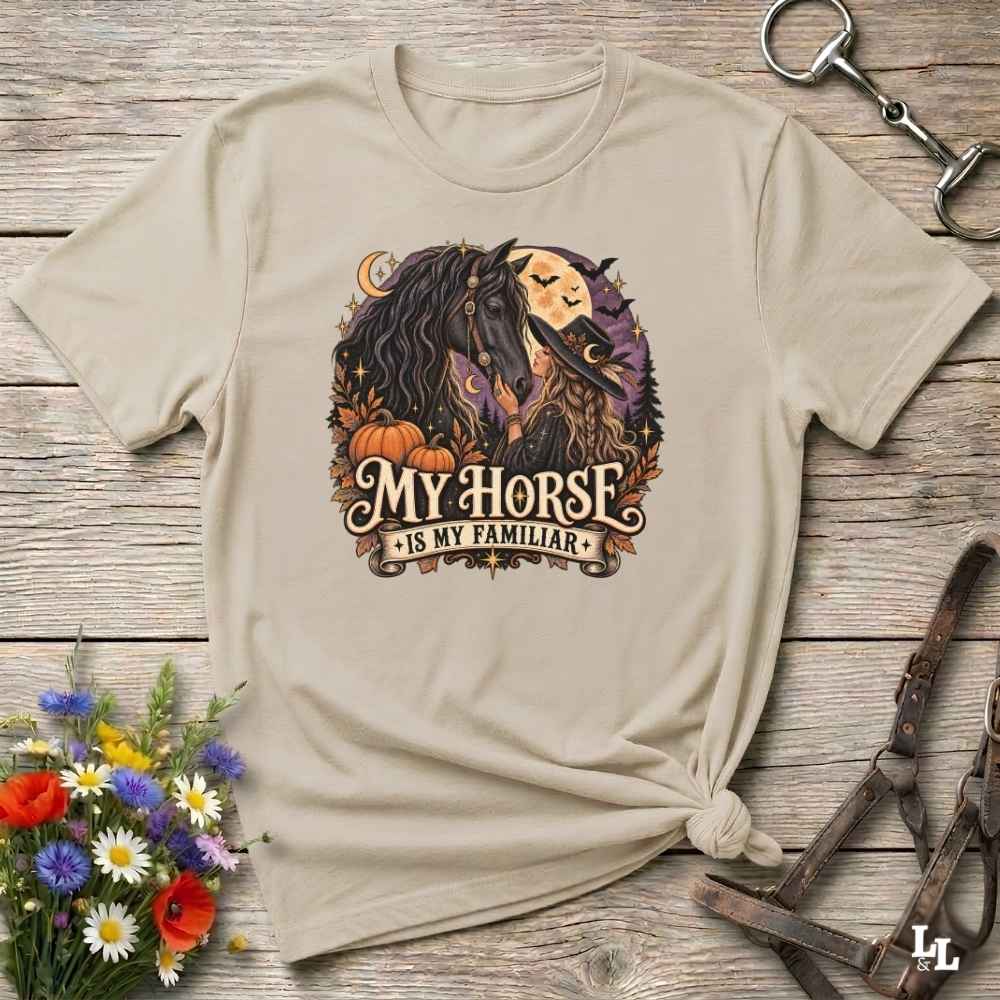 A beige graphic t-shirt with a 'My Horse Is My Familiar' design featuring a horse and witch illustration, styled flat on a rustic wooden background with a knot at the bottom.