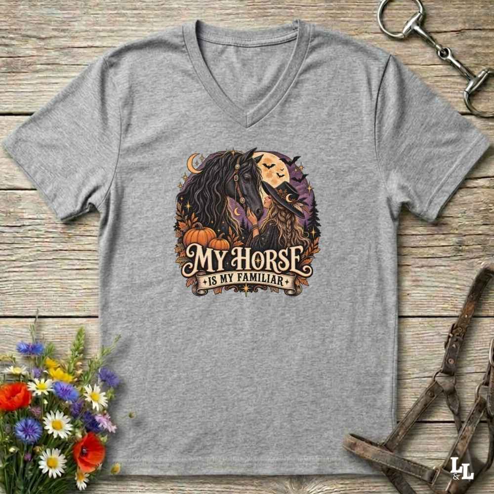 A heather gray V-neck t-shirt laid flat on a rustic wooden surface, featuring a circular graphic of a woman and horse with autumn elements and the text 'MY HORSE IS MY FAMILIAR'. Decorative flowers and equestrian tack are placed around the shirt.