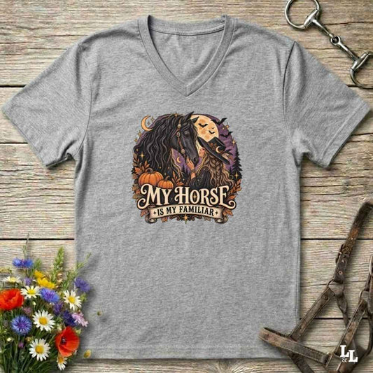 A heather gray V-neck t-shirt laid flat on a rustic wooden surface, featuring a circular graphic of a woman and horse with autumn elements and the text 'MY HORSE IS MY FAMILIAR'. Decorative flowers and equestrian tack are placed around the shirt.