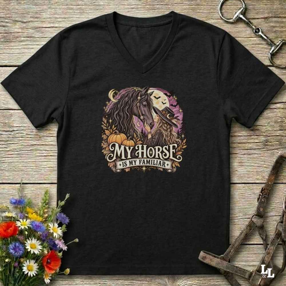 A black V-neck t-shirt laid flat on a rustic wooden surface, featuring a graphic of a horse and the text 'MY HORSE IS MY FAMILIAR' in a fall-themed design with pumpkins and leaves. The shirt is styled with a horse bit, halter, and wildflowers.