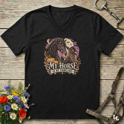 A black V-neck t-shirt laid flat on a rustic wooden surface, featuring a graphic of a horse and the text 'MY HORSE IS MY FAMILIAR' in a fall-themed design with pumpkins and leaves. The shirt is styled with a horse bit, halter, and wildflowers.