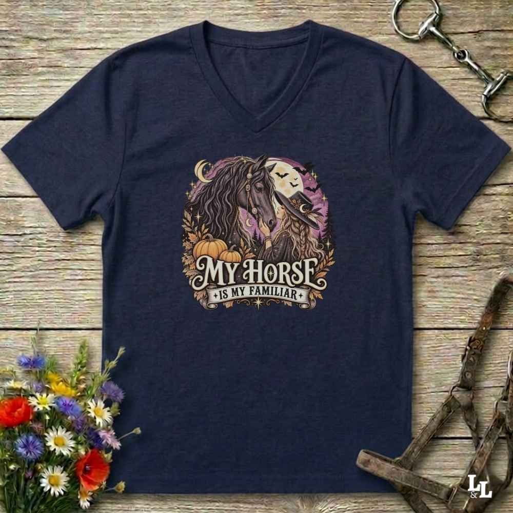 A navy blue V-neck t-shirt laid flat on a rustic wooden surface, featuring a graphic of a horse and the text 'My Horse Is My Familiar' in a circular design with pumpkins and autumn leaves. Decorative flowers and horse tack are placed around the shirt.