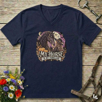 A navy blue V-neck t-shirt laid flat on a rustic wooden surface, featuring a graphic of a horse and the text 'My Horse Is My Familiar' in a circular design with pumpkins and autumn leaves. Decorative flowers and horse tack are placed around the shirt.