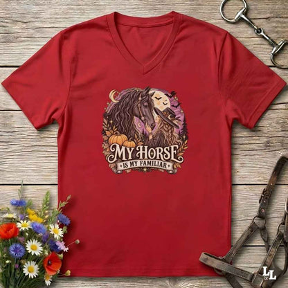 A red V-neck t-shirt laid flat on a rustic wooden surface, featuring a graphic of a horse and moon with the text 'MY HORSE IS MY FAMILIAR'. The shirt is styled with equestrian accessories and a small flower arrangement.